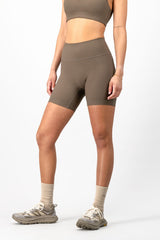 A model wearing high-waisted athletic biker shorts in a taupe brown color, paired with a matching sports bra, beige socks, and trail running shoes against a light gray studio background.